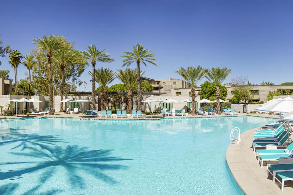 Resort pool with turquoise water, lounge chairs, palm trees, and modern white building with clear blue sky