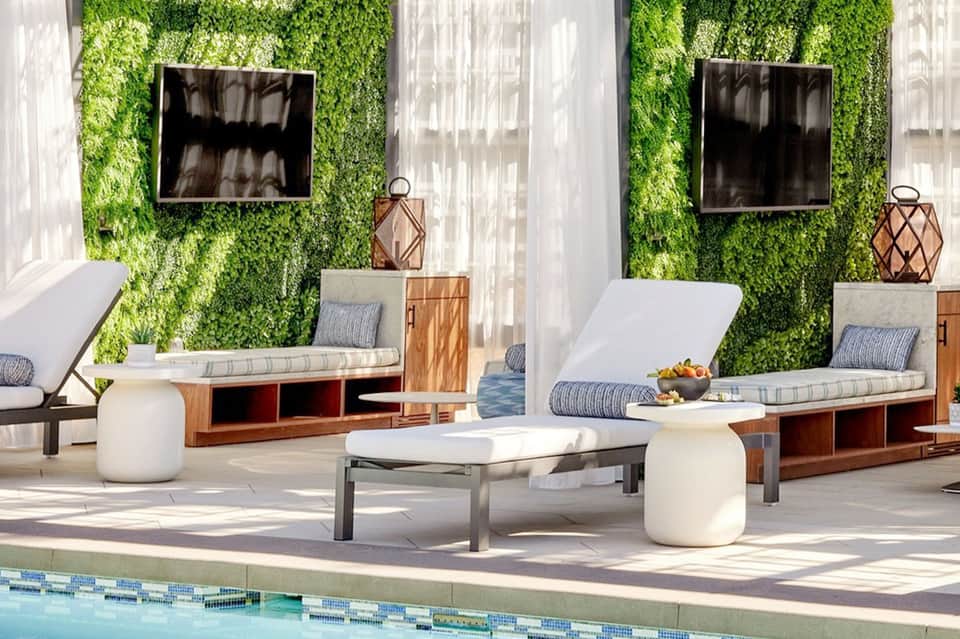 Poolside lounge area with white sun loungers, green moss walls, dark TVs, wooden storage units, and sheer curtains by the pool.