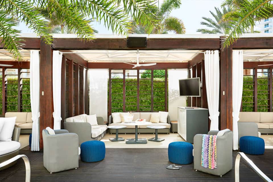 Covered outdoor lounge area with wooden beams, white curtains, cream seating, blue poufs, and palm trees overhead