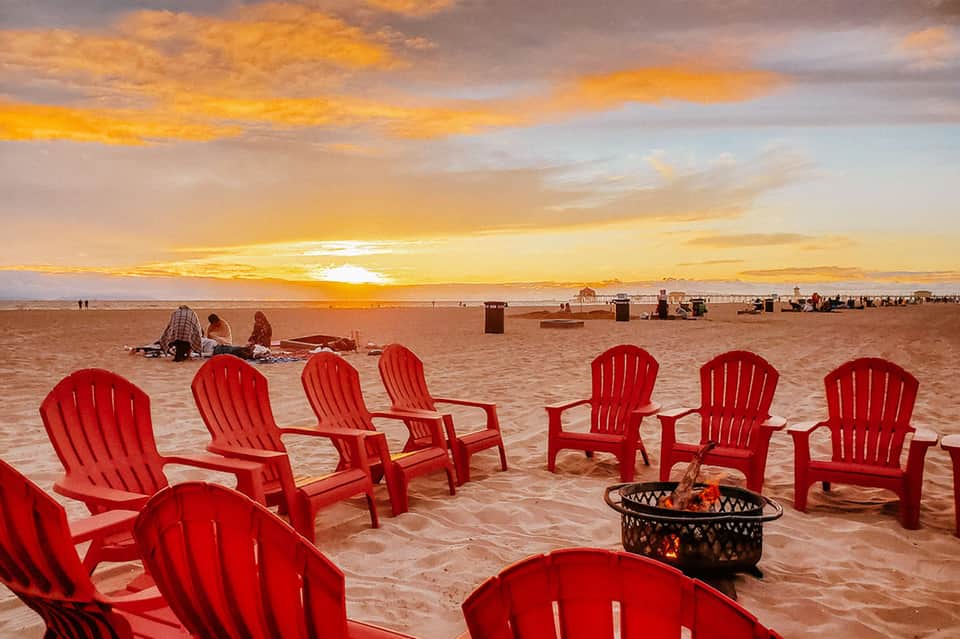 Red Adirondack chairs arranged on beach at sunset with fire pit