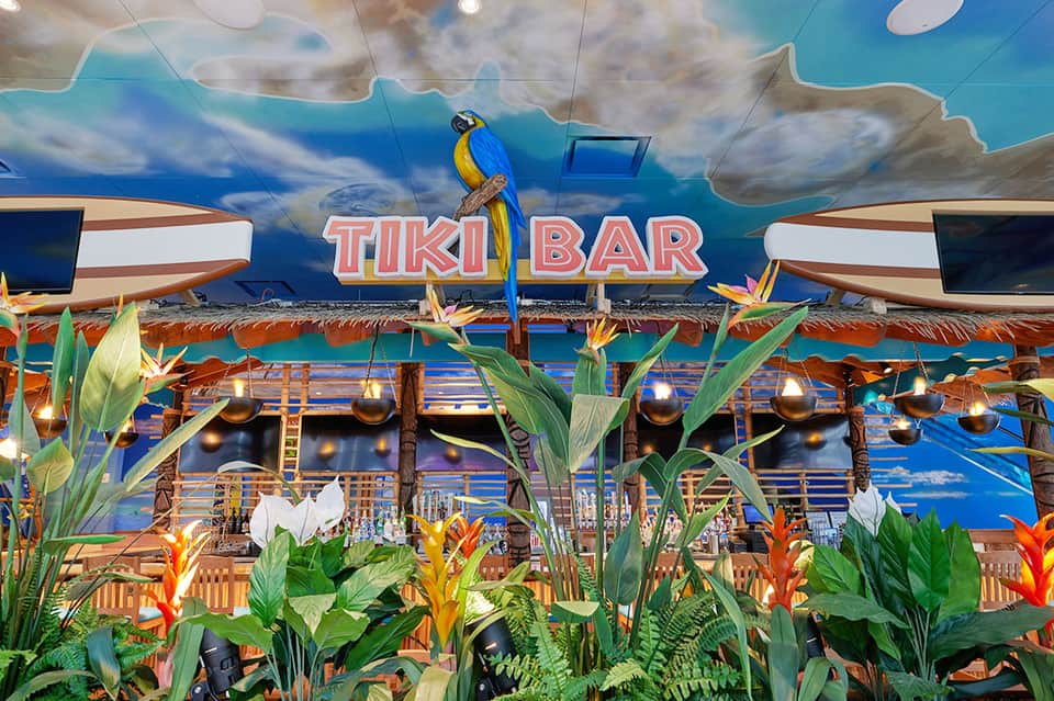 Tiki Bar tropical-themed indoor lounge with parrot statue, blue sky mural, and decorative flowers