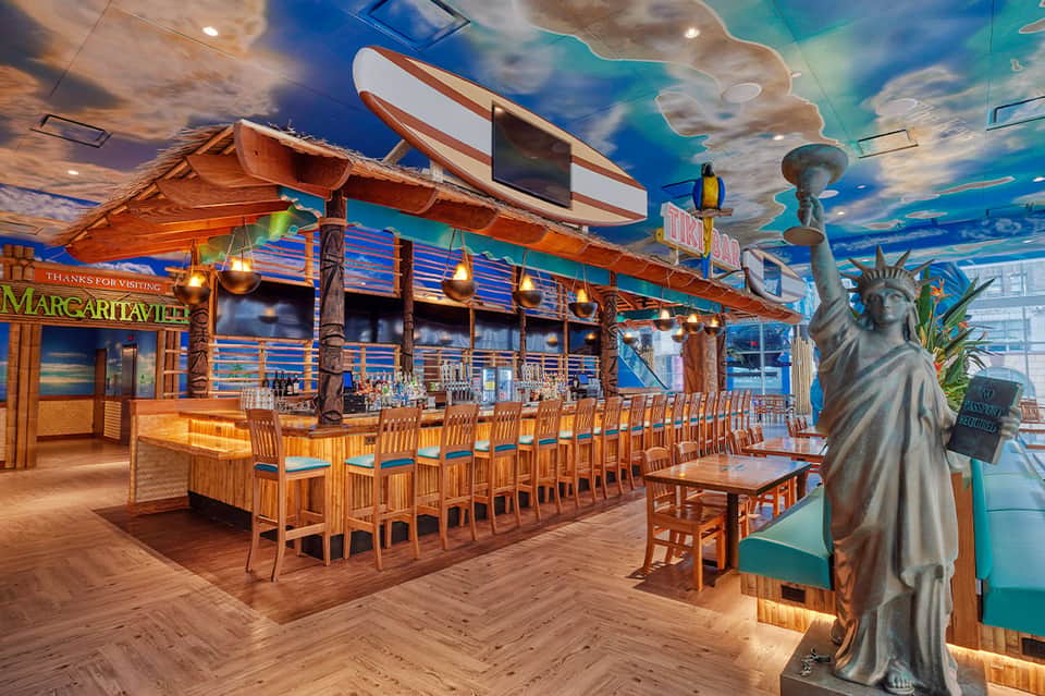 Tropical-themed bar restaurant interior with wooden tiki décor, ocean mural ceiling, and Statue of Liberty sculpture.