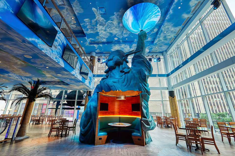 Indoor dining area with blue sculptural figure artwork and sky ceiling, surrounded by wooden chairs