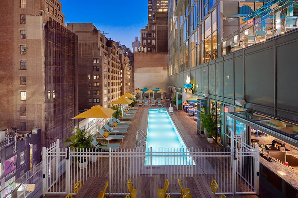 Rooftop pool deck in urban setting with lounge chairs, yellow umbrellas, and surrounding city buildings at dusk