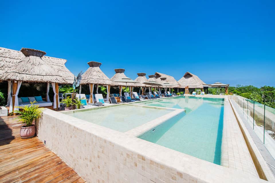 Luxury resort pool with thatched-roof cabanas, white loungers, and tropical landscaping under clear sky