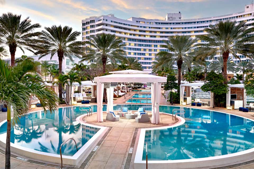Resort pool with white cabanas and lounge chairs surrounded by palm trees and high-rise hotel