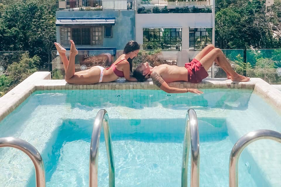 Two people relaxing on pool deck edge with modern architecture and lush greenery in background
