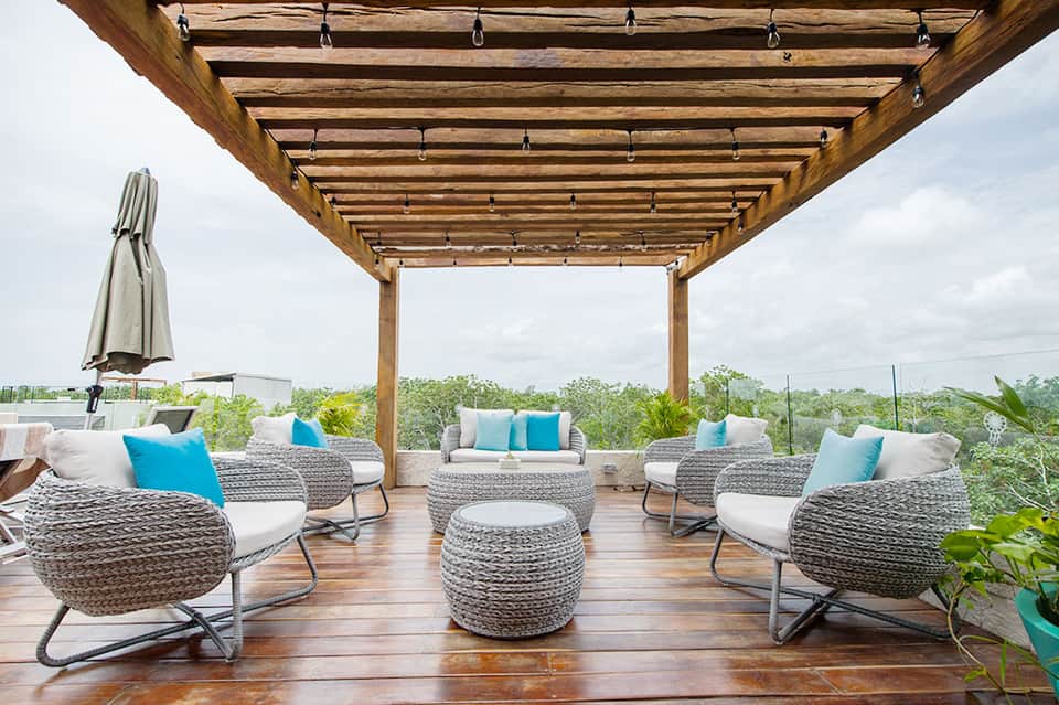 Modern rooftop lounge with woven chairs, blue pillows, wooden pergola, and landscape views