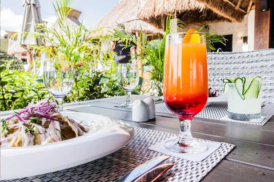 Tropical outdoor dining area with colorful cocktails, salad, and thatched-roof bungalows in background