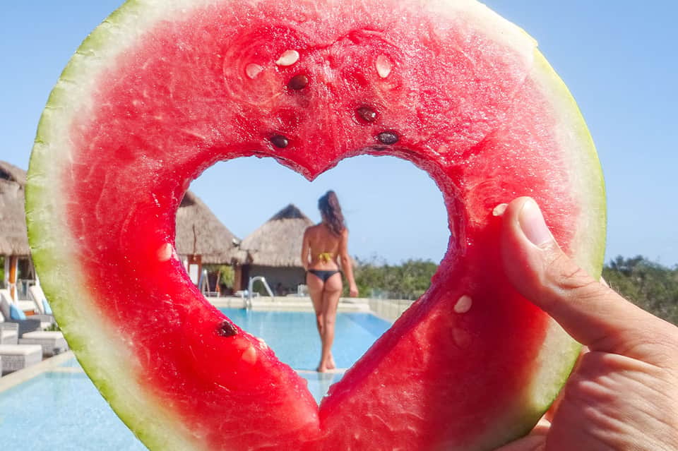 Heart-shaped watermelon slice held up with resort pool and palm-thatched cabanas in background
