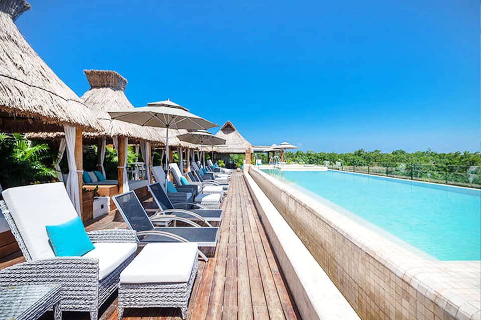 Resort pool deck with thatched cabanas, white lounge chairs, and turquoise infinity pool overlooking tropical landscape