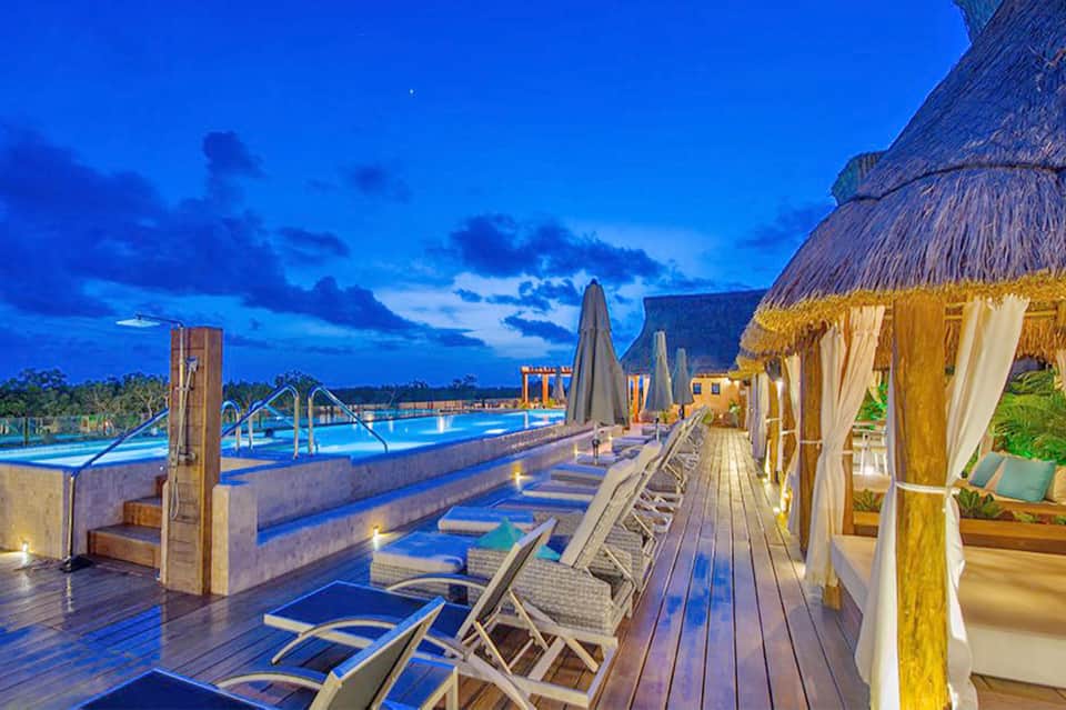 Illuminated resort pool deck at dusk with lounge chairs, umbrellas, and thatched-roof cabanas beside glowing pool