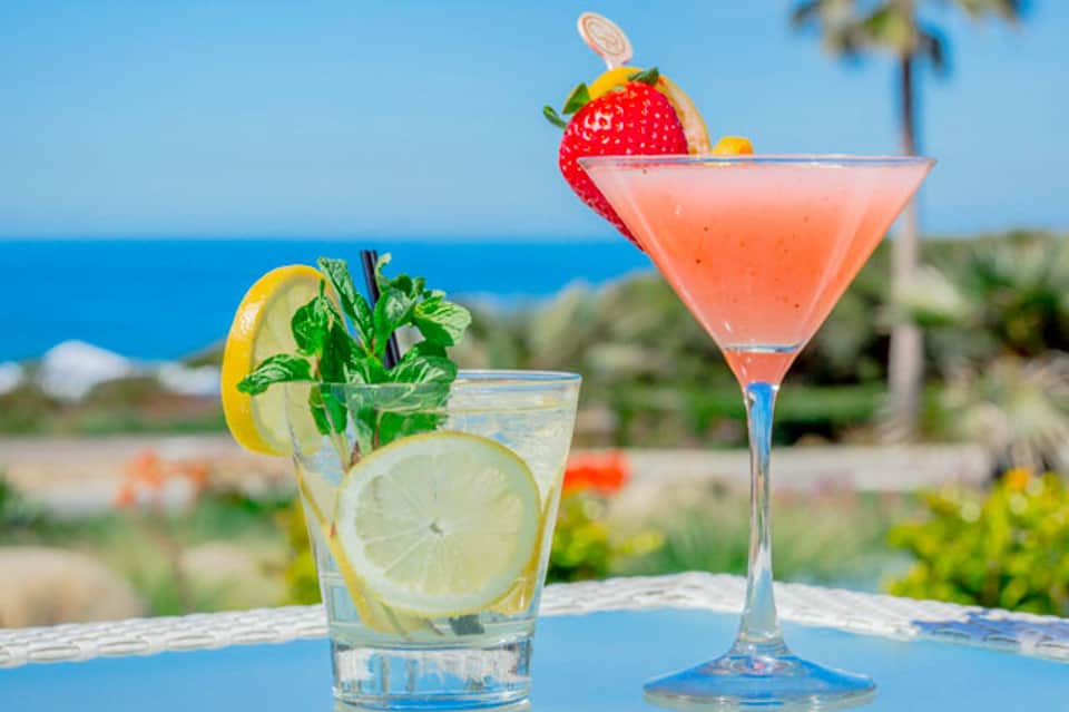Tropical cocktails with fresh fruit garnishes on a blue table overlooking the ocean and palm trees