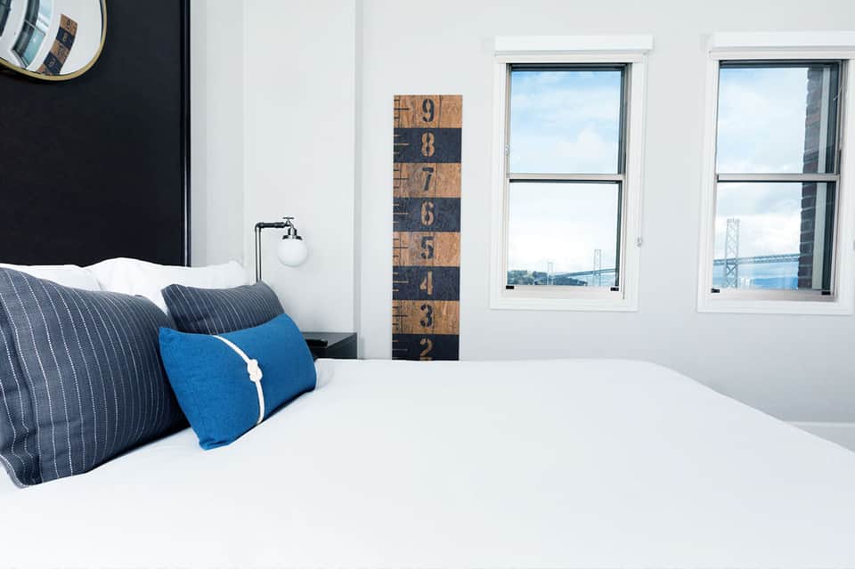 Modern hotel bedroom with white bedding, blue pillows, and bay bridge view through windows