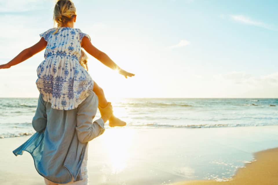 Adult holding child on shoulders at beach with arms outstretched toward ocean