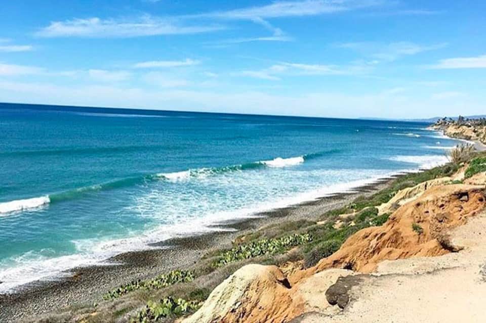 Coastal beach view with turquoise ocean, white sand, and eroded cliffs under blue sky