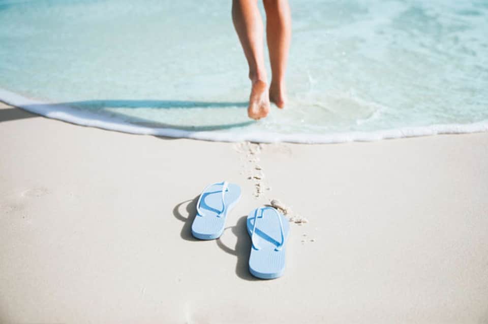 Pair of blue flip-flops on white sand beach with ocean waves and surfboard in background