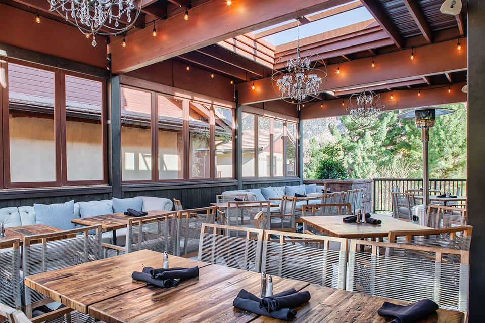 Modern covered patio dining area with wooden tables, chairs, and crystal chandeliers overlooking green landscape