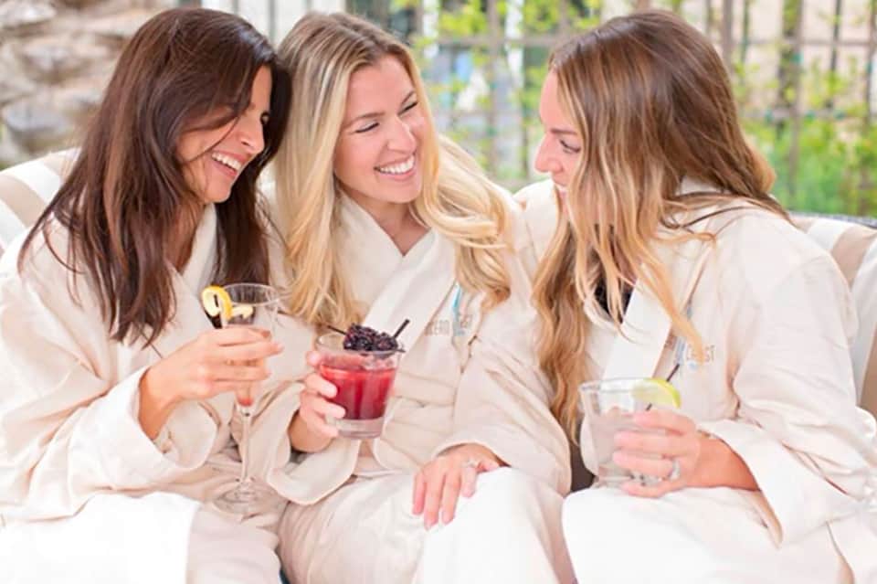 Three women in white robes toasting with cocktails and beverages indoors