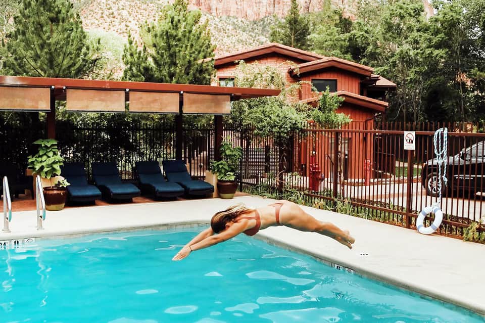 Person diving into a bright blue pool at a mountain resort with red lodge buildings and forest backdrop