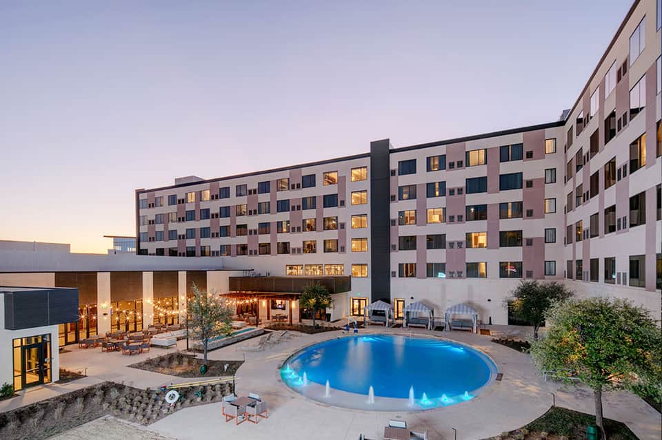 Resort courtyard at dusk with illuminated pool, multi-story hotel building, and lounge seating