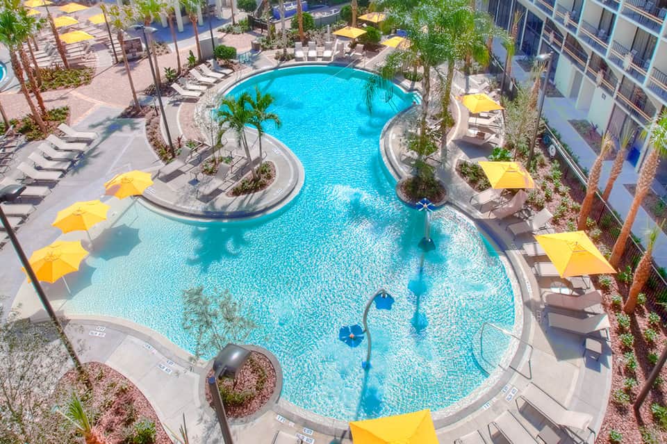 Aerial view of curved resort pool with yellow umbrellas, palm trees, and surrounding buildings