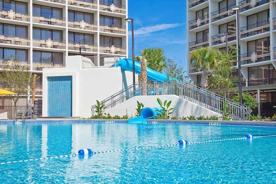 Resort pool with blue water slide and modern white building with balconies under clear sky