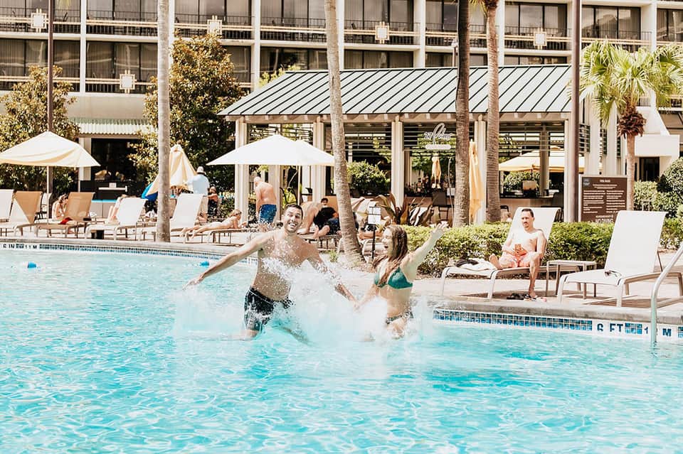 Family enjoying resort pool with modern hotel building, white umbrellas, and palm trees in background