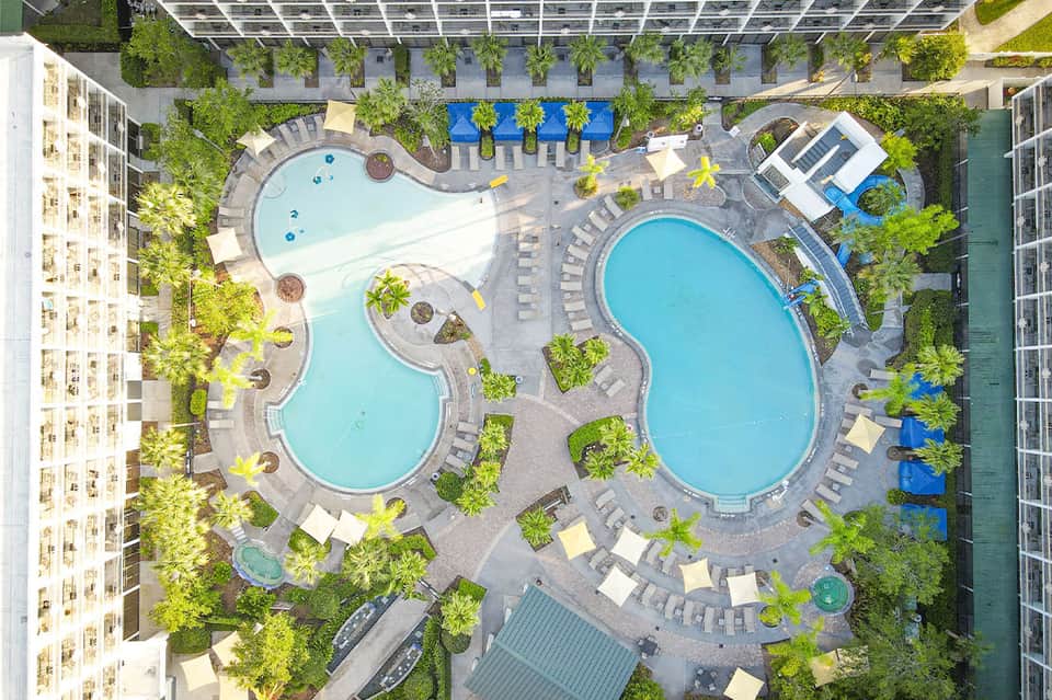Aerial view of two resort pools surrounded by lounge chairs, tropical plants, and buildings