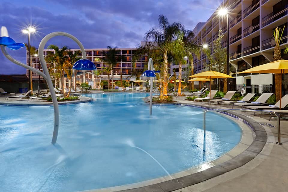 Resort pool at dusk with blue umbrellas, palm trees, and multi-story hotel buildings lit by evening lights