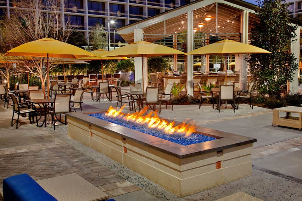 Illuminated fire pit on patio with yellow umbrellas, seating areas, and modern covered pavilion at dusk
