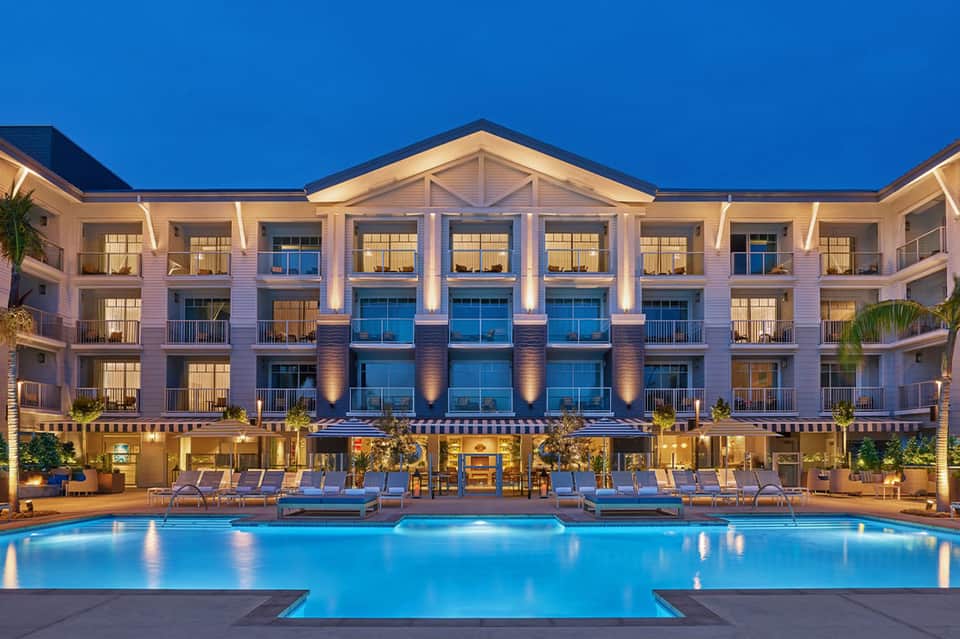 Modern resort exterior at dusk with illuminated pool, multi-story building, and loungers reflected in calm water