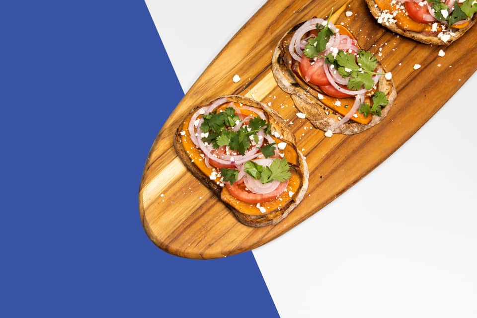 Three gourmet toasts with fresh toppings on wooden serving board against blue background