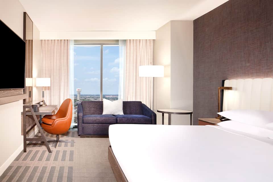 Modern hotel suite with bed, work desk, sofa, floor-to-ceiling windows overlooking cityscape