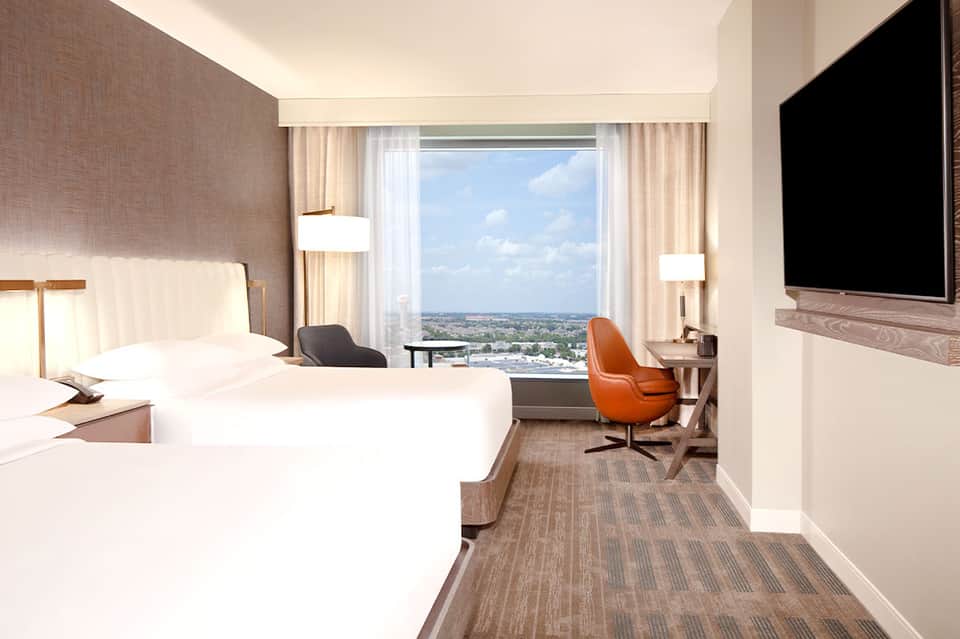 Spacious hotel room with queen beds, floor-to-ceiling windows overlooking landscape, orange accent chair, and flat-screen TV