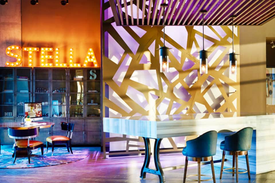 Modern hotel lobby with glowing Stella sign, geometric gold wall art, blue accent chairs, and ambient purple lighting