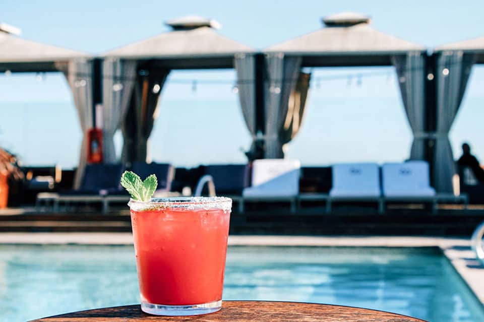 Red cocktail with mint garnish on poolside table, cabanas visible in background
