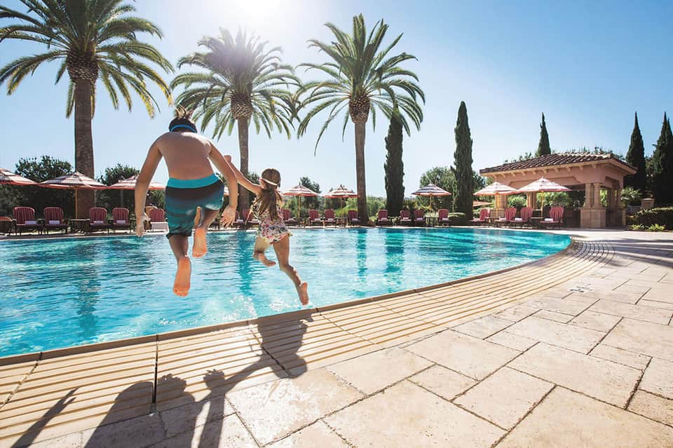 Father and child running into resort pool with palm trees and lounge chairs under Mediterranean sun
