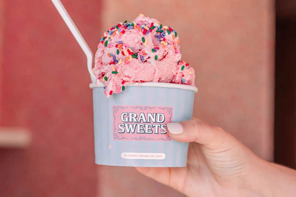 Pink ice cream with sprinkles in Grand Sweets branded cup held by hand