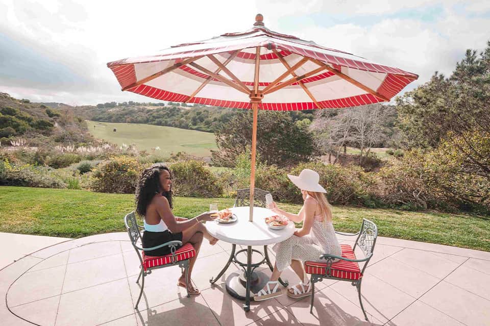 Two women dining under red and white striped umbrella at round table with scenic hillside valley backdrop