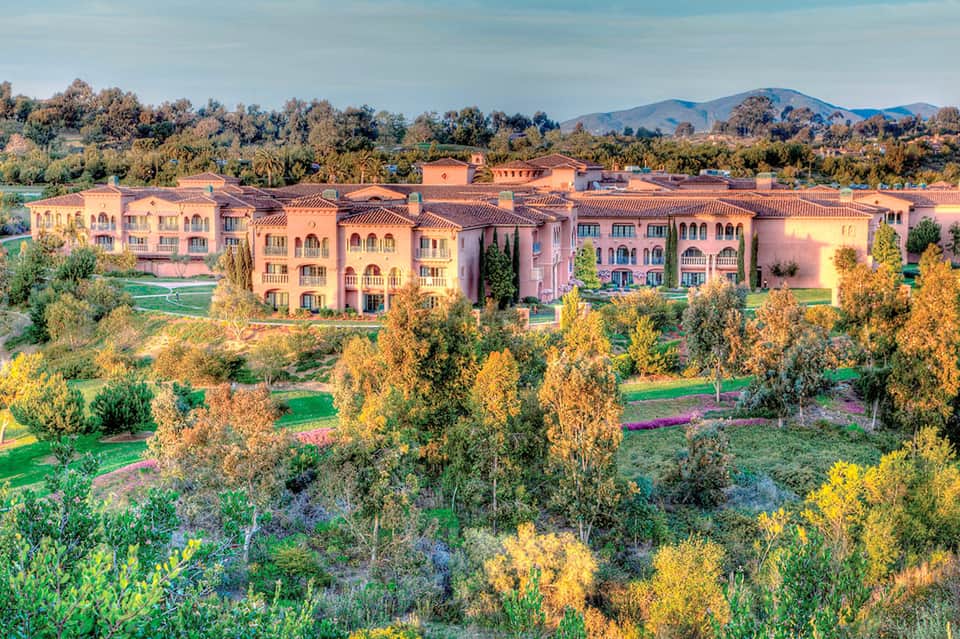 Aerial view of sprawling pink Mediterranean-style resort with manicured grounds and mountains in distance