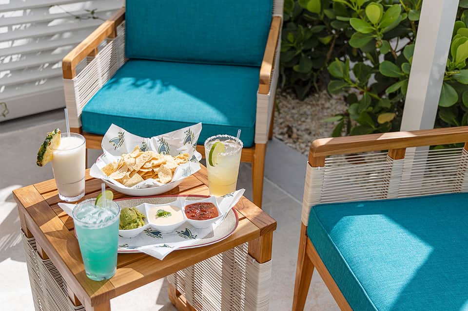 Beachfront patio seating with turquoise cushioned chairs, appetizers, and tropical greenery