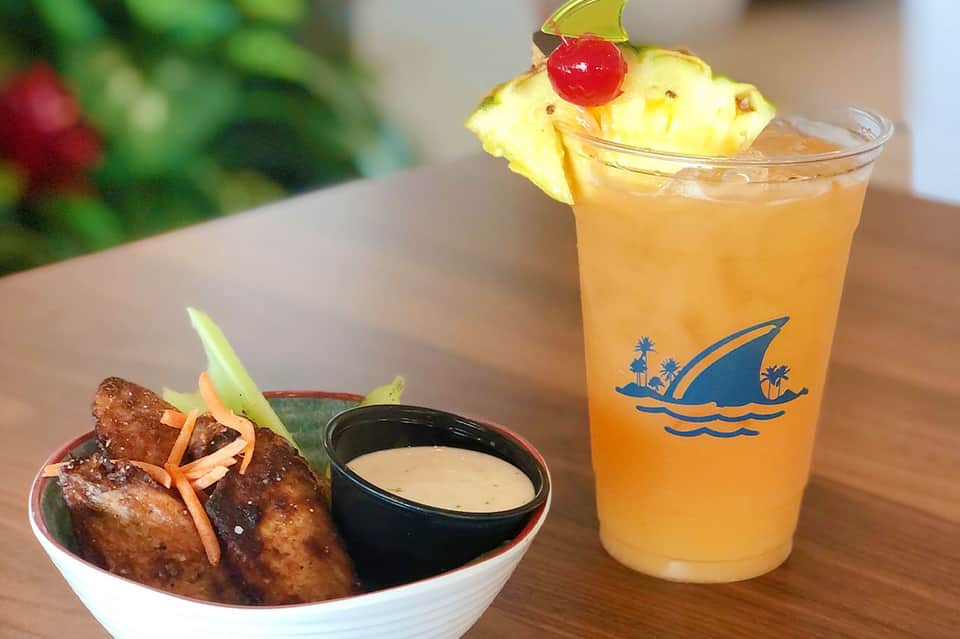 Tropical cocktail with pineapple garnish beside appetizer bowl with dipping sauce on wooden table