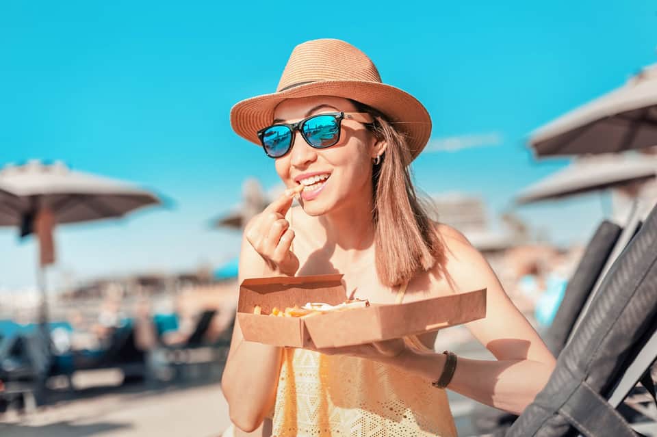 Woman in summer hat and sunglasses enjoying pizza at a beach resort with umbrellas and clear blue sky