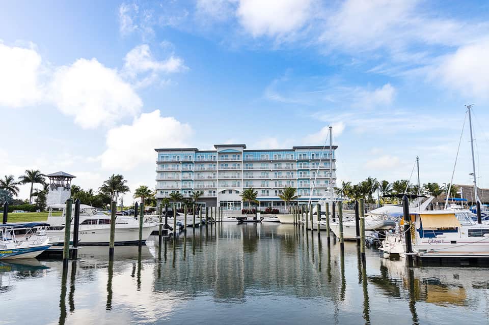 Waterfront hotel building with marina docks, boats, and palm trees under blue sky