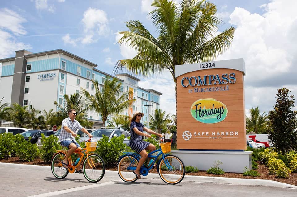 Two cyclists riding colorful bikes in front of Compass by Margaritaville hotel with palm trees