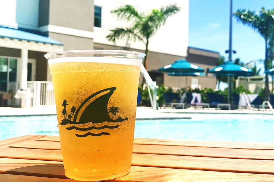 Yellow beverage with shark logo design in cup by resort pool with palm trees and blue umbrellas