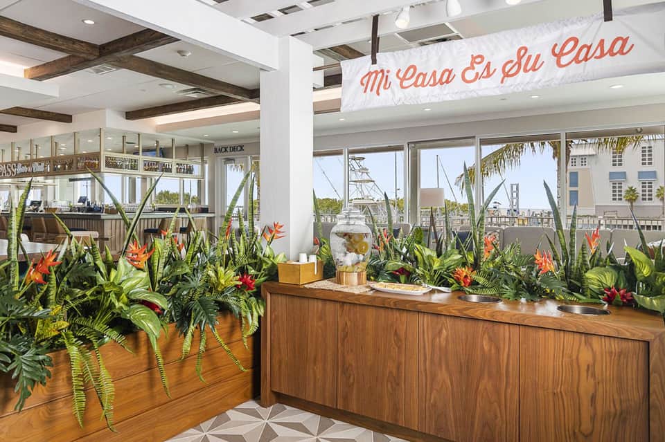Bright reception area with tropical plant arrangements and 'Mi Casa Es Su Casa' banner