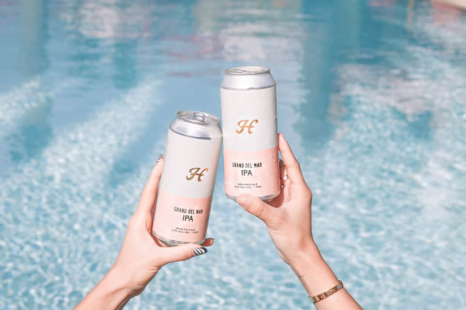 Two cans of Grand del Mar IPA beer held above a sparkling swimming pool.
