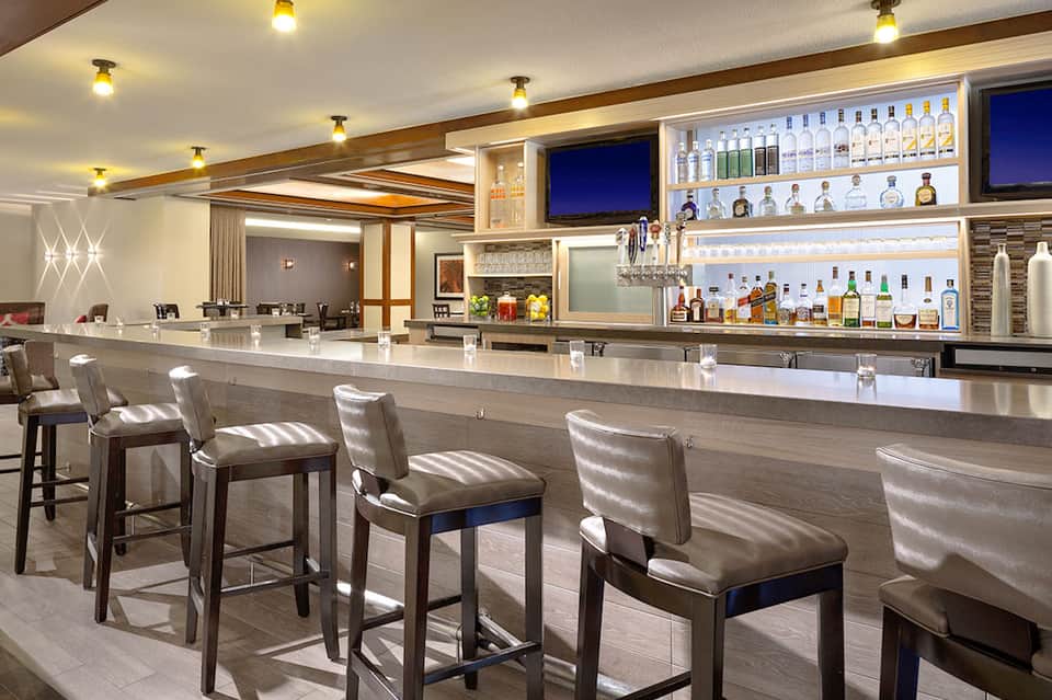 Modern hotel bar with white countertop, bar seating, and illuminated liquor shelves